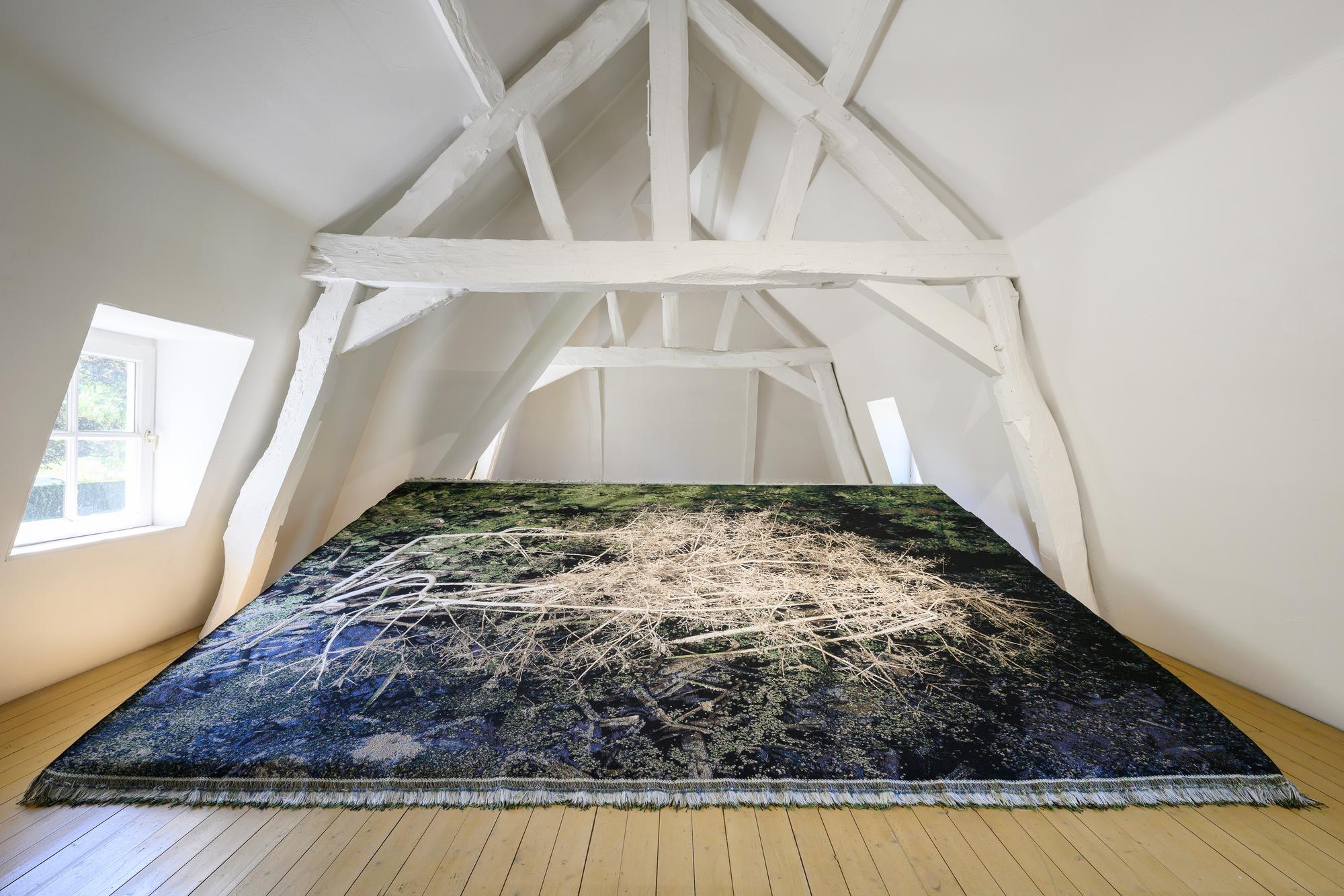 Grote textiele print van een wateroppervlak met takken ligt uitgespreid op de vloer van een zolderruimte met wit houten balkenplafond.