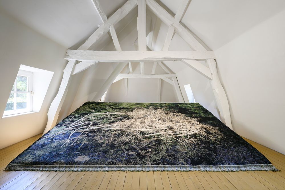 Grote textiele print van een wateroppervlak met takken ligt uitgespreid op de vloer van een zolderruimte met wit houten balkenplafond.