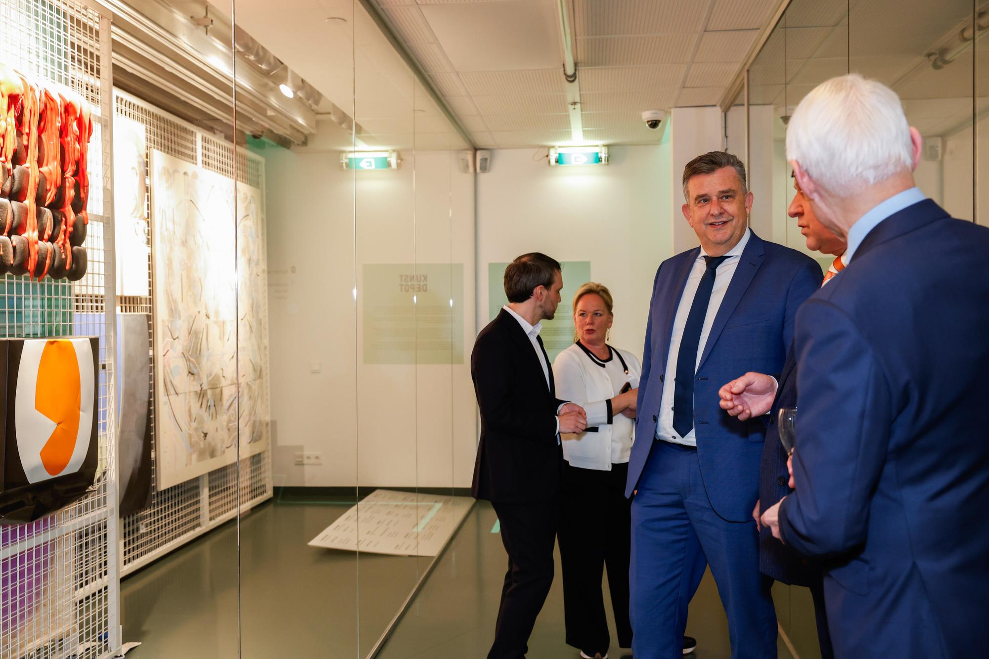 Twee mannen en een vrouw praten met elkaar terwijl ze in het Kunsdepot staan. Links is een rek met schilderijen te zien.