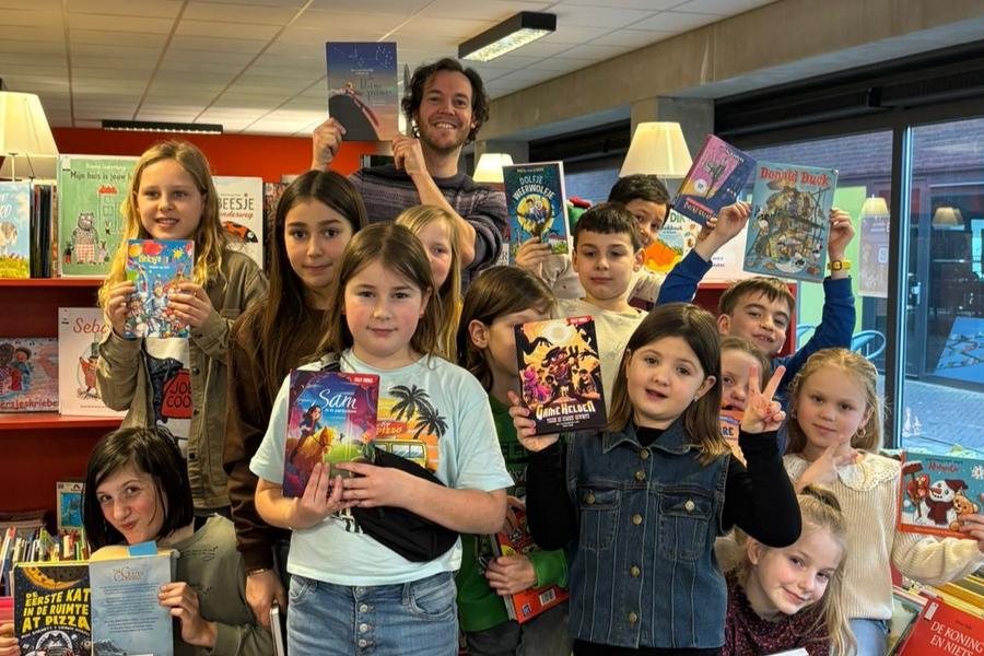Groep kinderen in een boekwinkel houdt verschillende kleurrijke kinderboeken omhoog, omringd door volle boekenrekken.