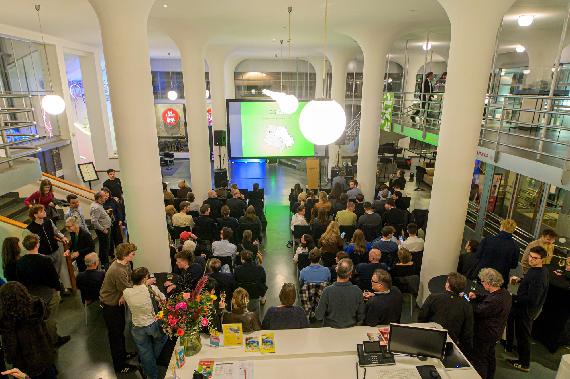 Bezoekers vullen het atrium van SCHUNCK tijdens de feestelijke uitreiking van de Euregional Architecture Prize 2025, met zicht op het podium en presentatiescherm.