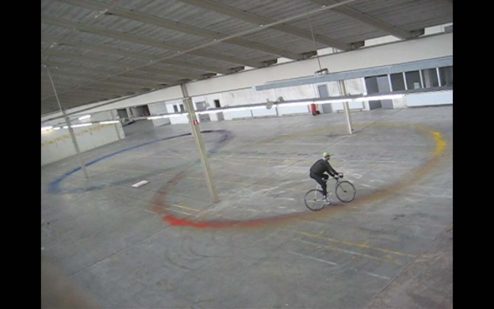 Een fietser rijdt in cirkels door een lege industriële hal, waarbij gekleurde verfsporen (rood, geel en blauw) op de vloer achterblijven. De persoon draagt een zwarte outfit en een groene helm.