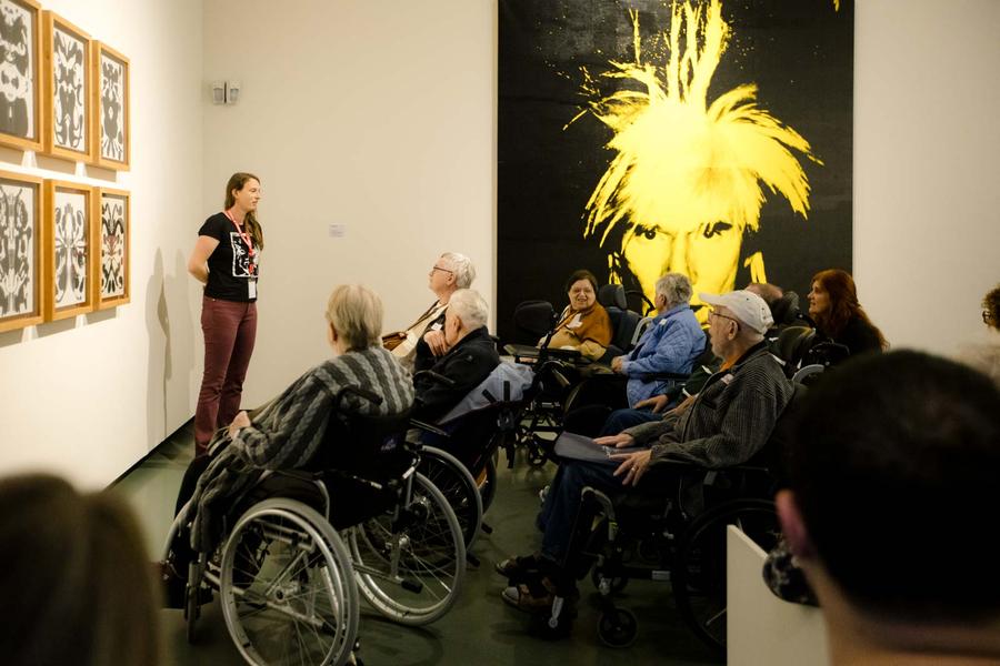 Bezoekers in rolstoelen luisteren naar rondleider in het museum tijdens Andy Warhol Vanitas. 