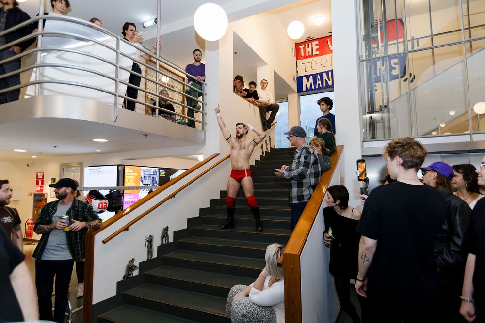 Een performer in rode sportbroek en zwarte laarzen komt energiek de trappen af in een moderne, lichte hal, omringd door toeschouwers op de trap en balustrade; mensen kijken, lachen en filmen terwijl de man met beide armen omhoog wijst, onder ronde lampen en kleurrijke posters aan de muur. Tijdens Frictions Festival #10 bij SCHUNCK Glaspaleis. 