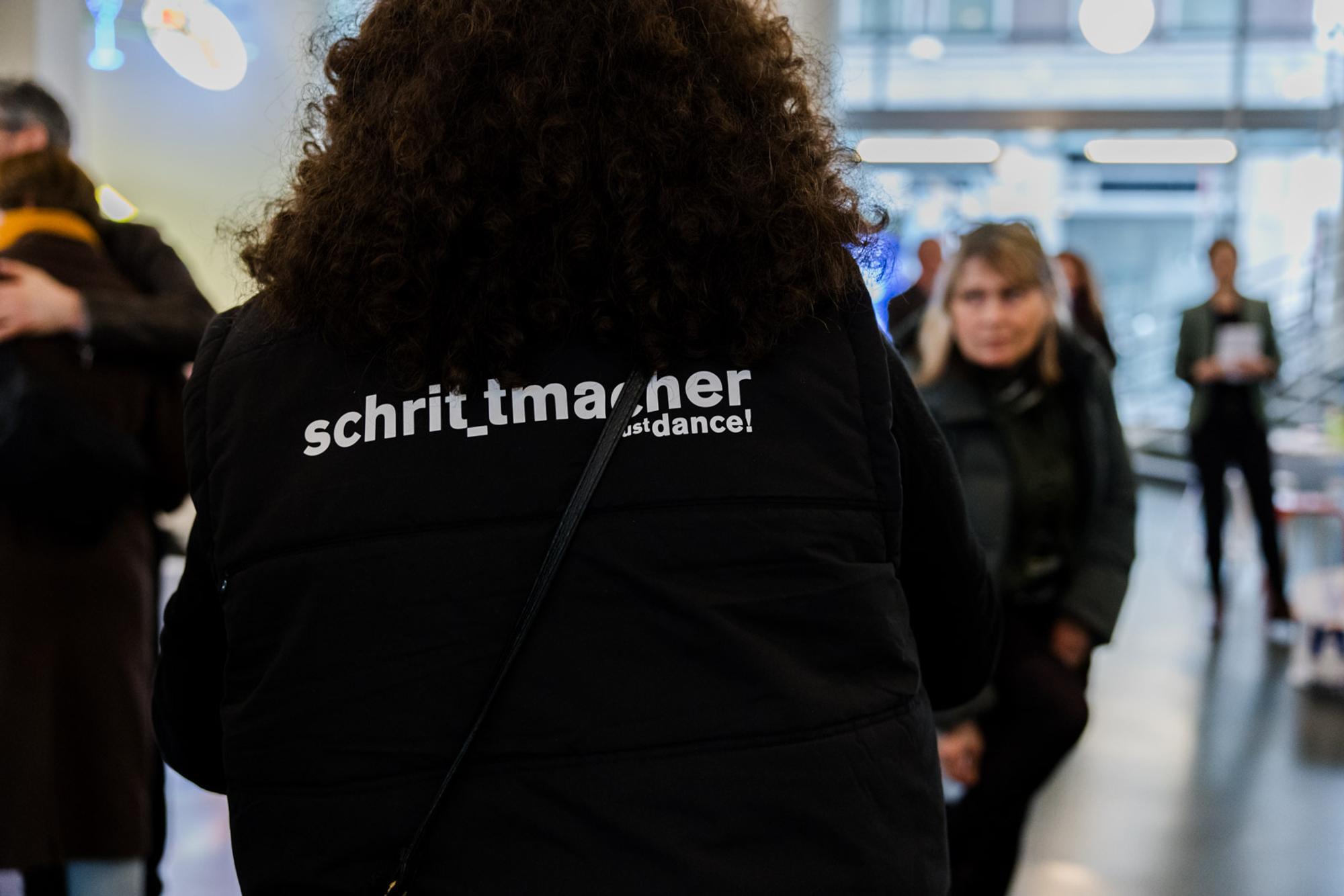 Persoon met zwarte bodywarmer van de achterkant, met in witte letters op de rug: "schrit_macher, just dance!" Op de achtergrond bezoekers in de hal van SCHUNCK.