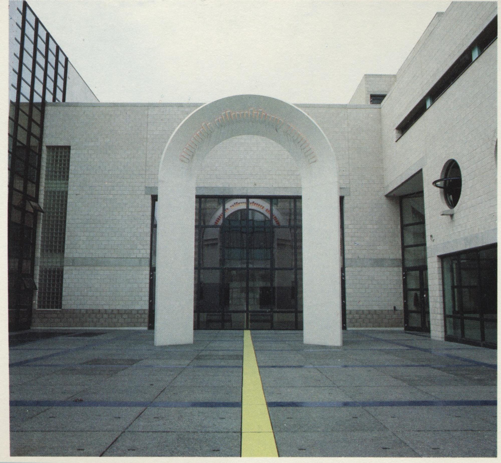 Moderne gebouwentree met een grote witte boog en een gele lijn op het plein die naar de glazen ingang leidt.
