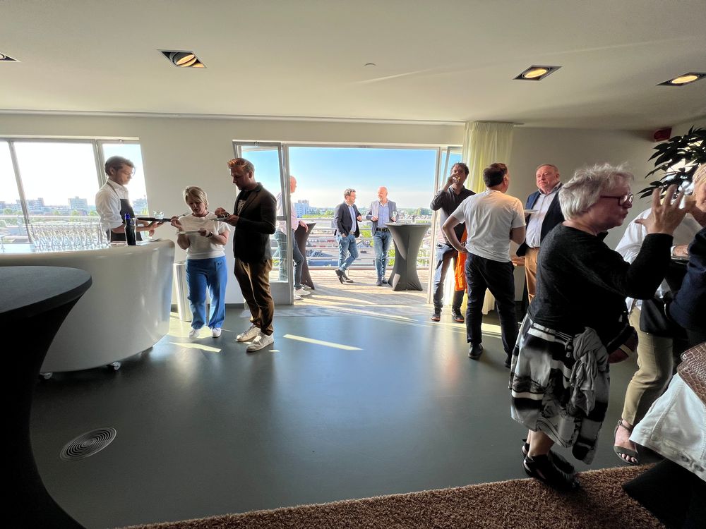 Een groep mensen staat en praat in een lichte ruimte met grote ramen en uitzicht op de stad; links wordt drank geserveerd aan een ronde witte bar, terwijl enkele gasten op het balkon genieten van het uitzicht.