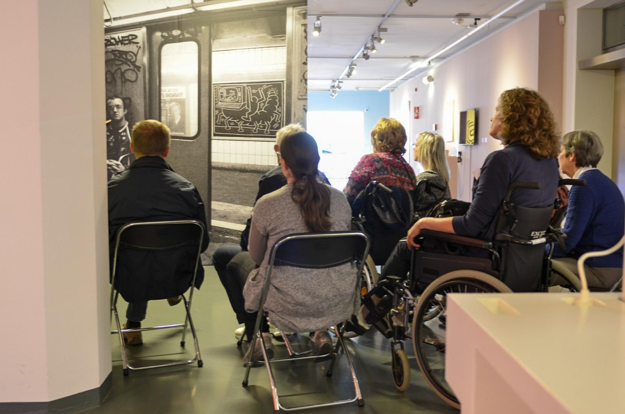 Een groep mensen zit op stoelen, gericht naar een muur met een grote zwart-witfoto van wat lijkt op een metrotrein. Eén persoon zit in een rolstoel. De setting lijkt een binnenruimte te zijn, mogelijk een kunstgalerij of museum, met bovenverlichting en enkele kunstwerken die aan de muren zijn tentoongesteld.