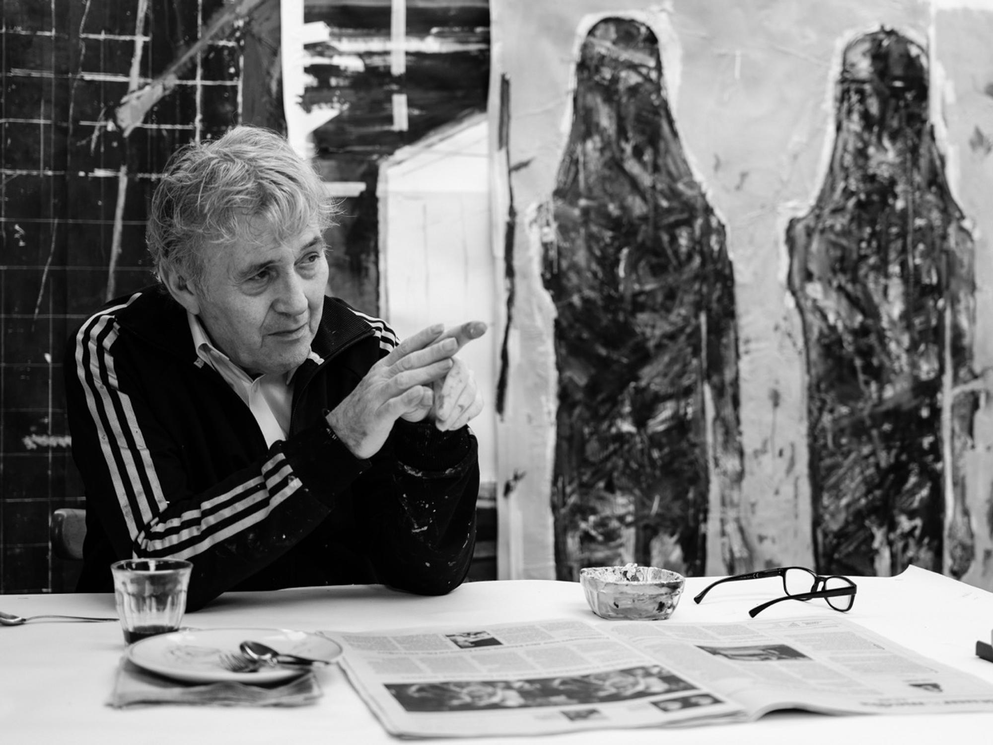 André Joosten zit links achter een tafel en wijst. rechts van hem zie je een kunstwerk. Op de tafel liggen een krant een bril een bord en een glas