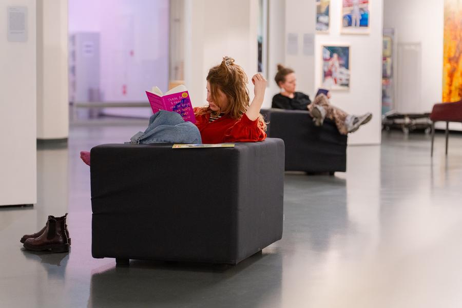 Twee personen zitten in comfortabele stoelen in een moderne kunstgalerie; één leest een felroze boek, de ander gebruikt een smartphone. Op de vloer staat een paar laarzen.