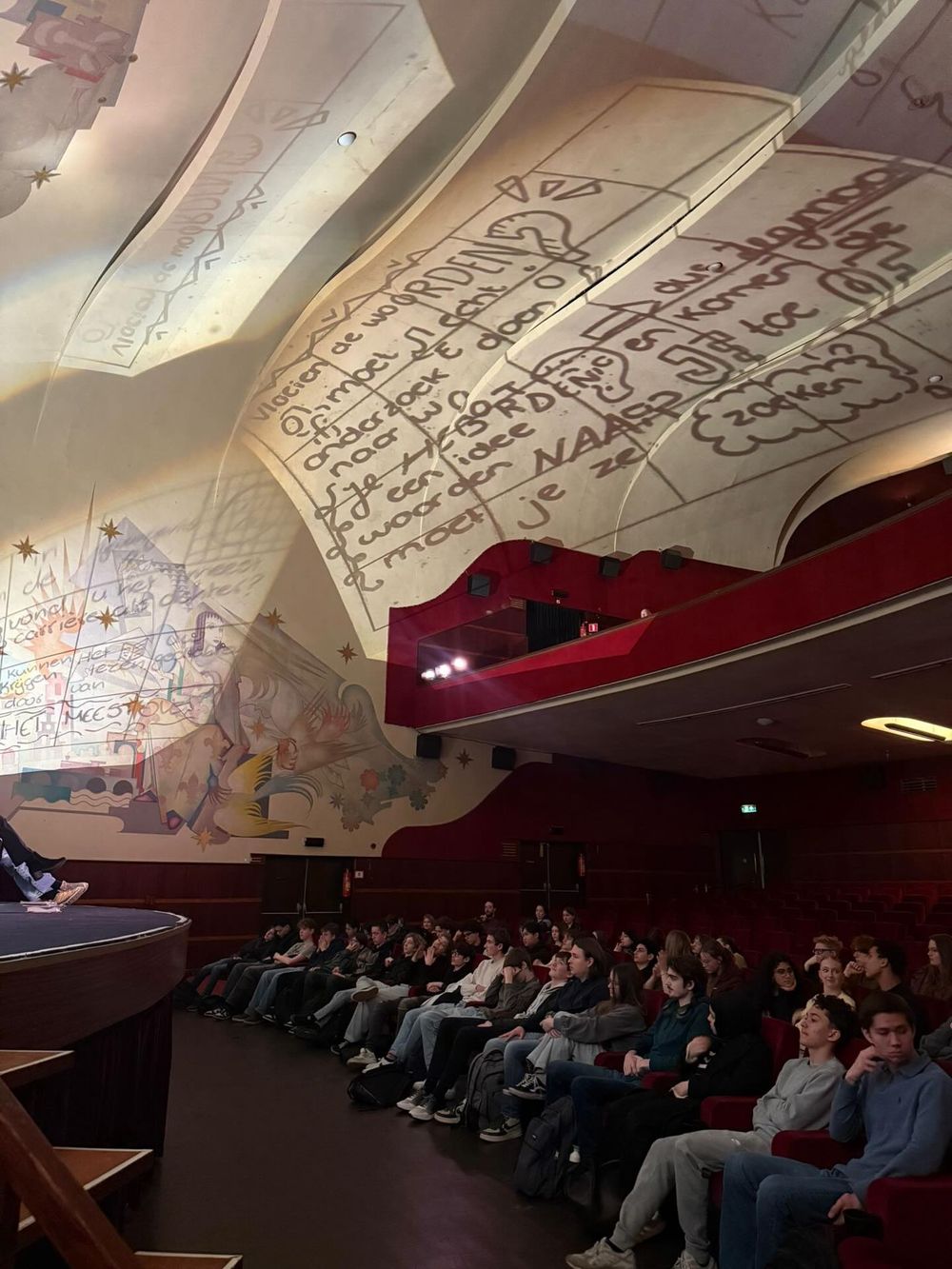 Publiek in een theaterzaal kijkt naar een dichter op het podium terwijl handgeschreven poëzie groot op het plafond wordt geprojecteerd.