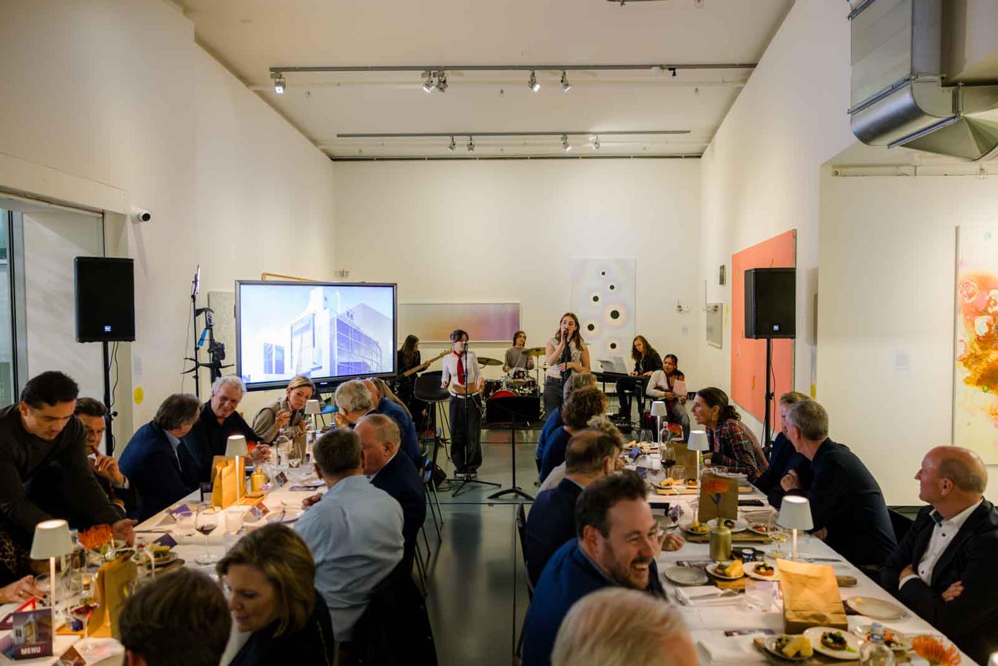 Mensen aan lange tafels in het museum terwijl jongeren een muzikaal optreden verzorgen.