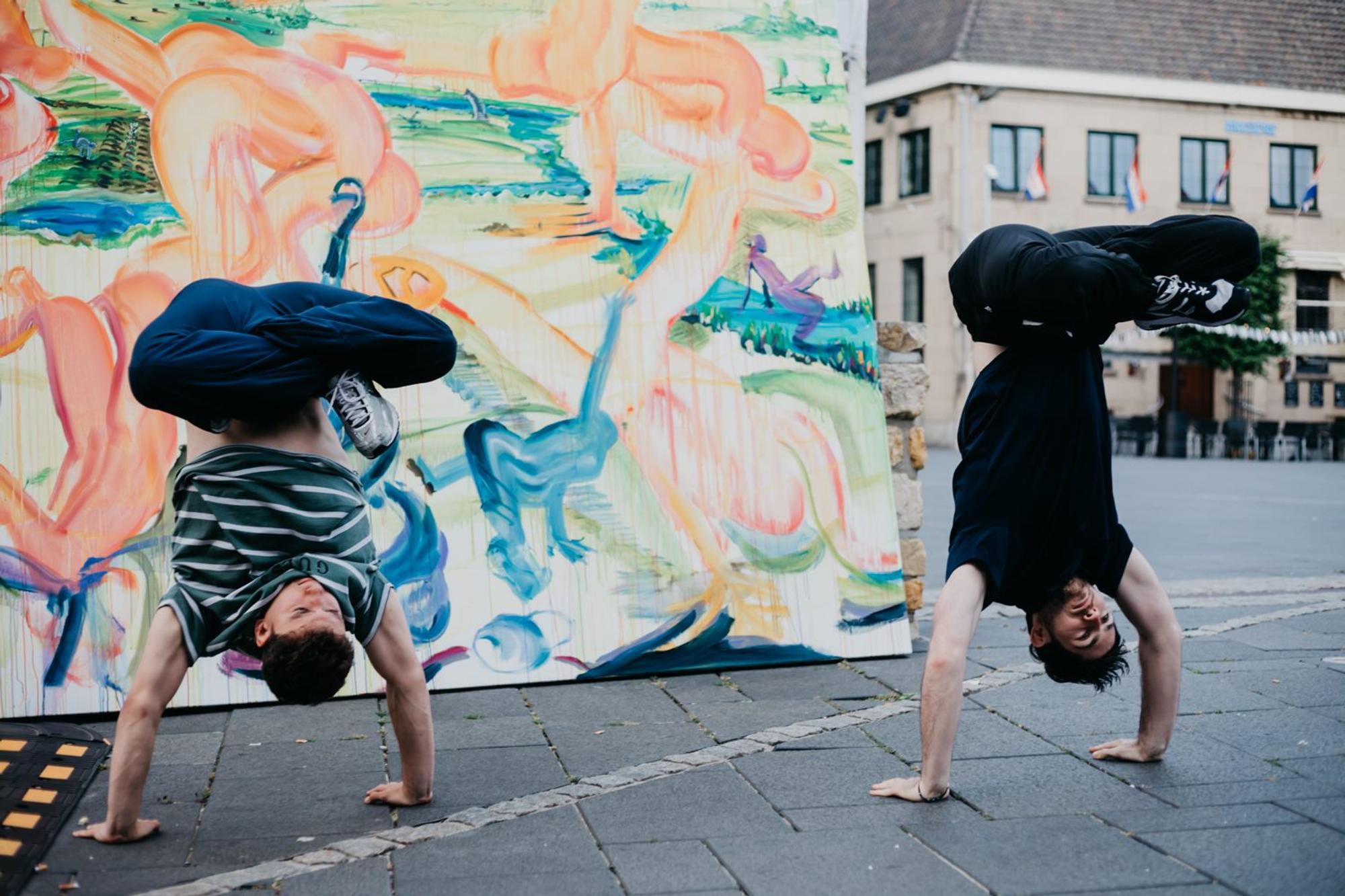 Twee jongens die breakdansen staan buiten voor een schilderij waarop dansende figuren te zien zijn, op hun handen in kleermakerszit met hun benen in de lucht.