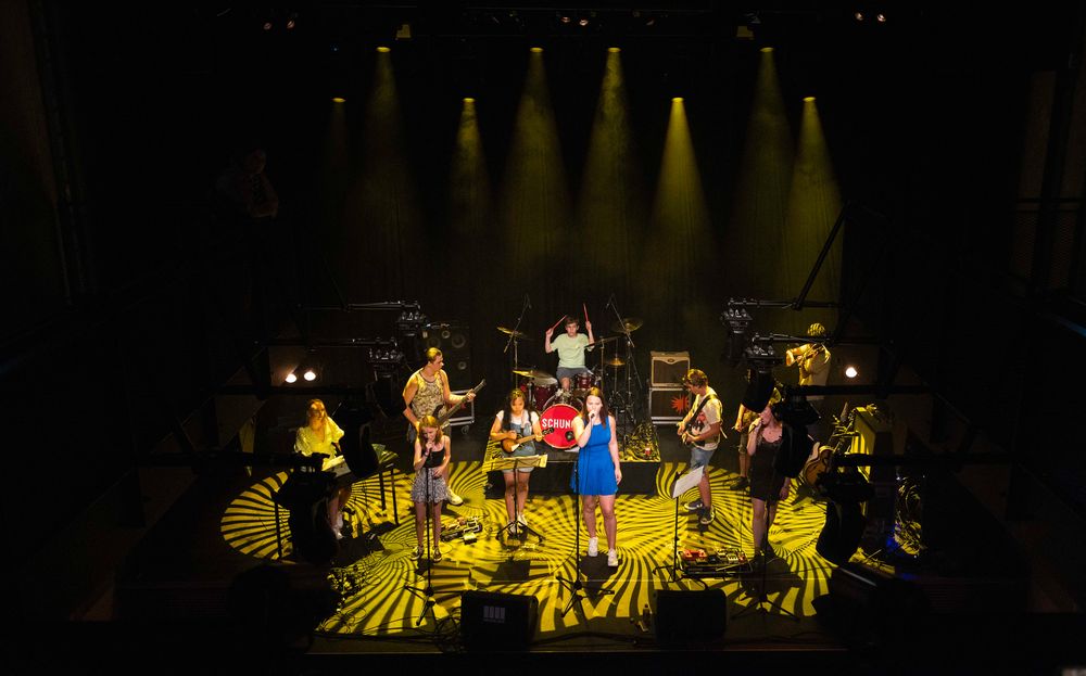 Op een podium tijdens een optreden van SCHUNCK Academy staat een volledige bandopstelling met meerdere muzikanten verspreid over het podium. In het midden staat een zanger bij een microfoon, met daaromheen gitaristen en toetsenisten, terwijl achteraan een drummer op een verhoogd platform speelt onder geel podiumlicht en patronen op de vloer.
