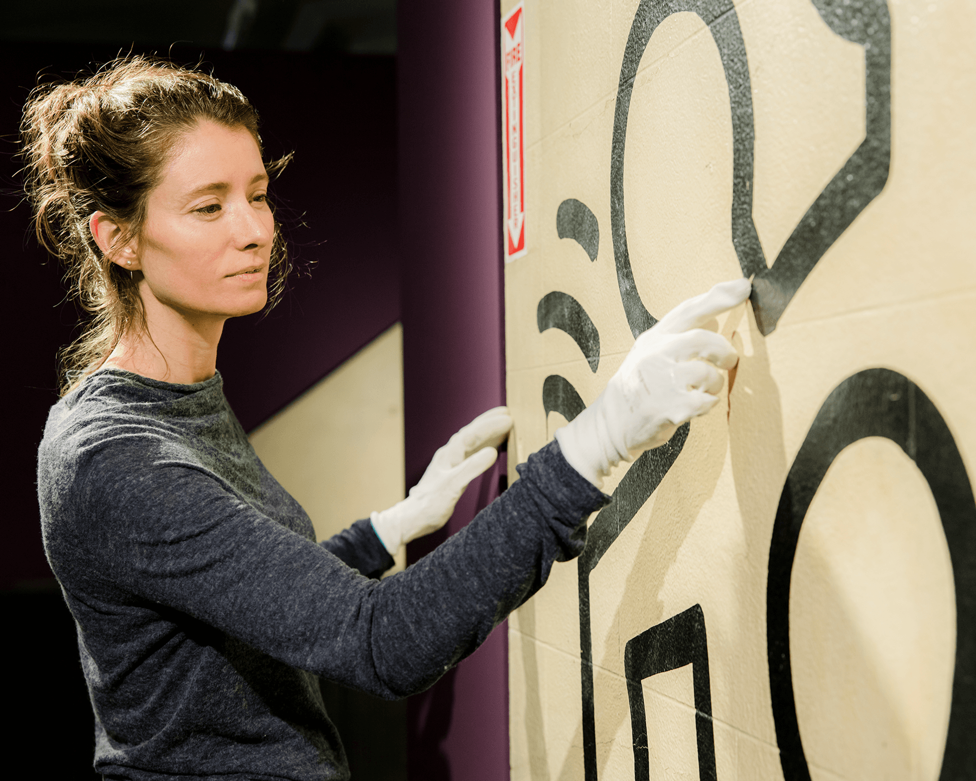 Een persoon met witte handschoenen wijst naar een zwart ontwerp op een muur. 