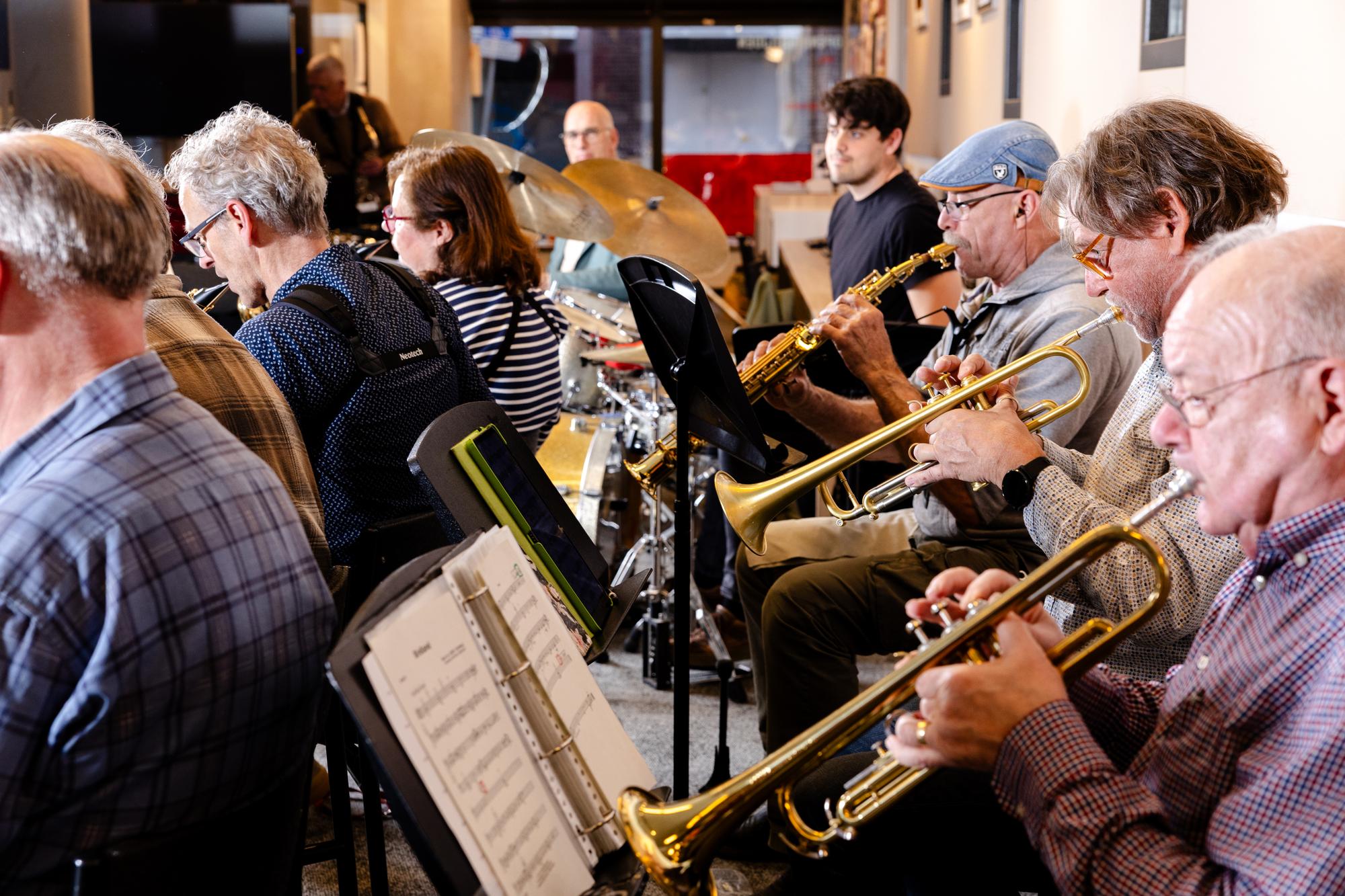Volwassen muzikanten spelen samen tijdens een orkestrepetitie, met trompetten, saxofoons en slagwerk. Op de voorgrond zijn bladmuziekstandaards zichtbaar, terwijl de groep geconcentreerd musiceert in een informele repetitieruimte.
