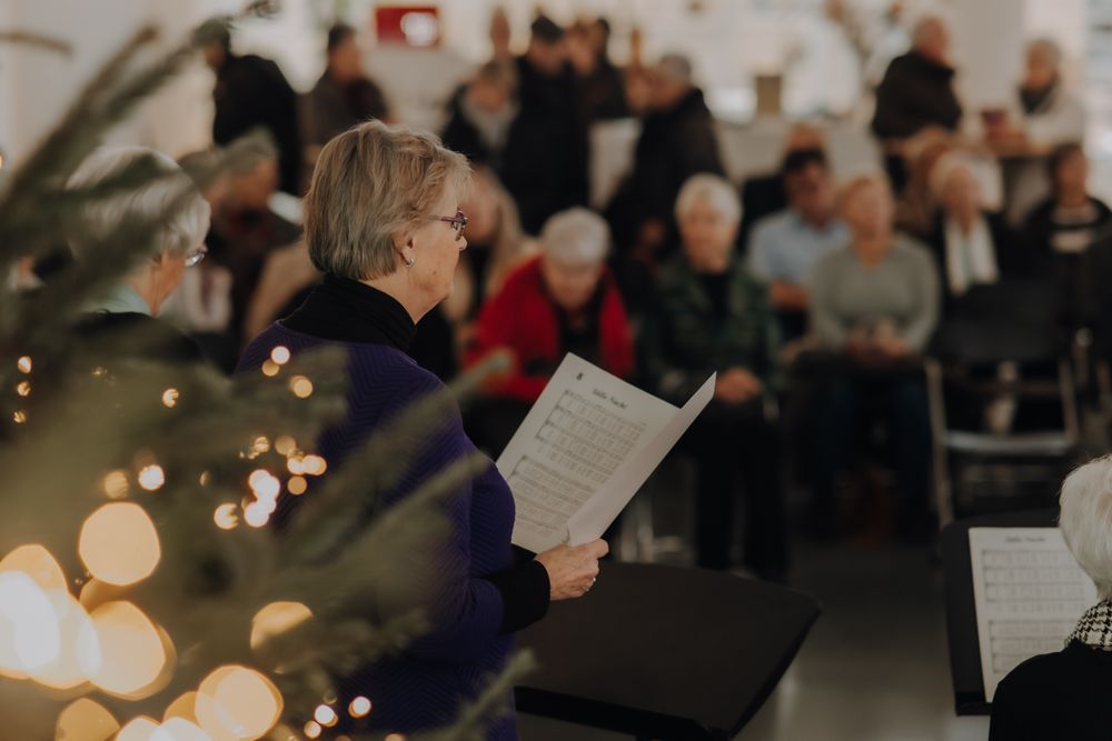 Vrouw houdt bladmuziek vast en zingt in het koor tijdens een winterconcert. Het publiek is vaag zichtbaar op de achtergrond, zittend op stoelen, terwijl een kerstboom met lichtjes subtiel op de voorgrond zichtbaar is.