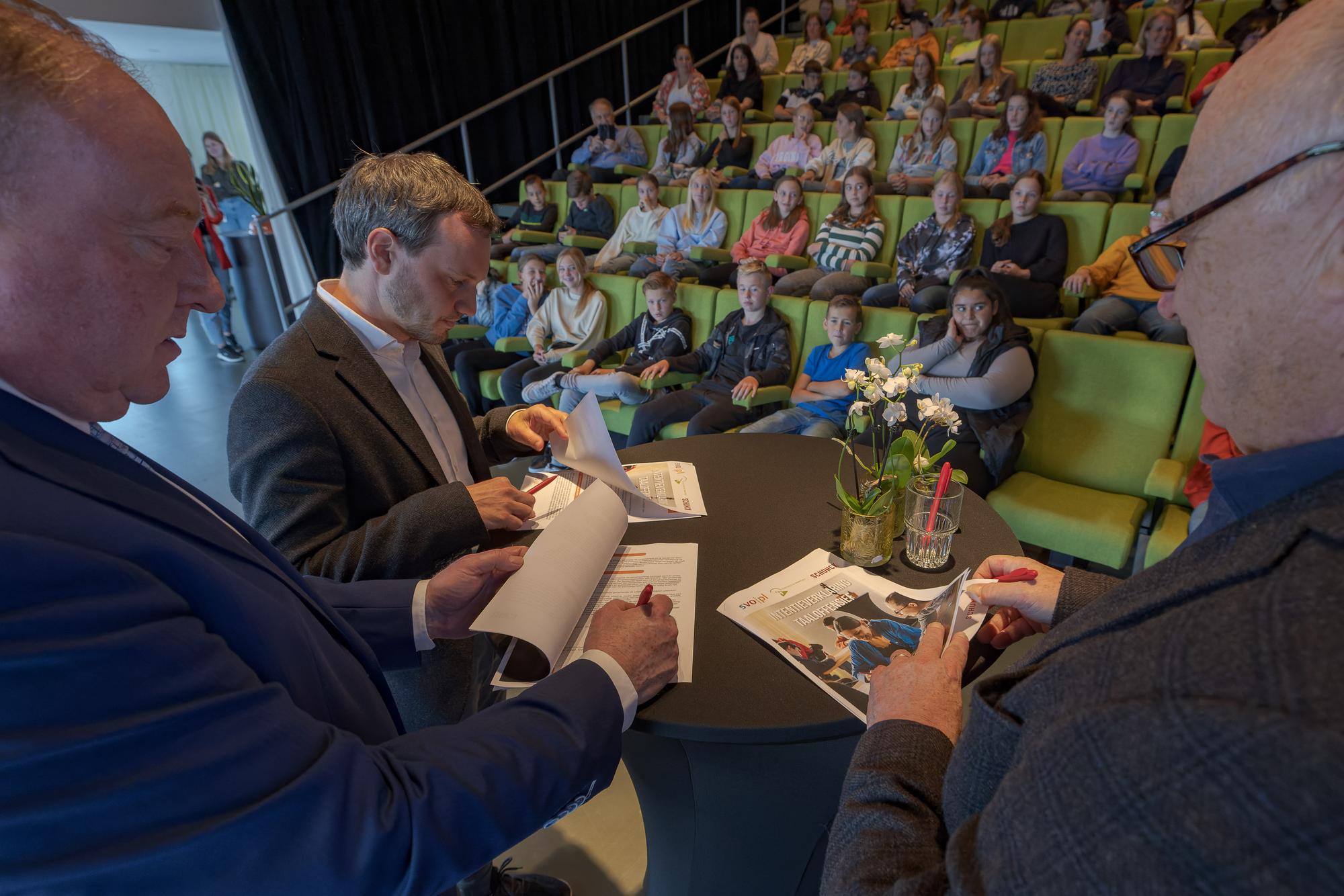 Drie mannen staan om een statafel met papieren op tafel in het auditorium van SCHUNCK. Op de achtergrond is een tribune met bezoekers te zien.