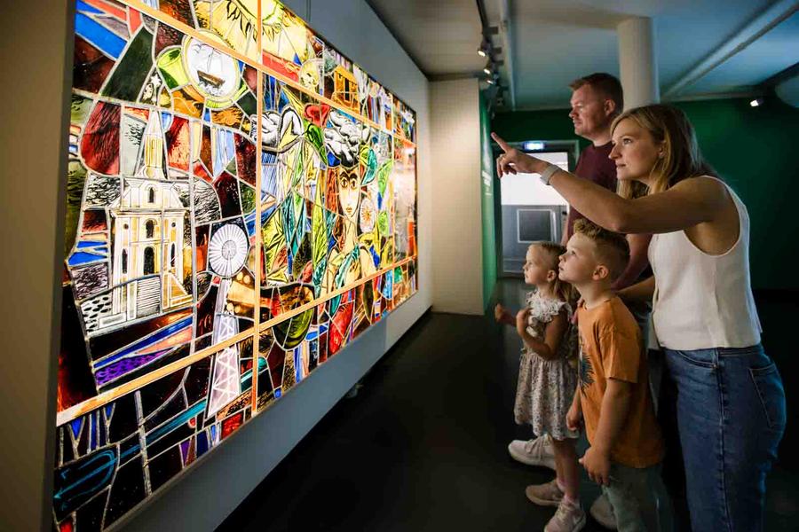 Een gezin met twee jonge kinderen bekijkt samen een kleurrijk kunstwerk van glas-in-lood in SCHUNCK Glaspaleis. De moeder wijst iets aan terwijl de kinderen aandachtig toekijken.