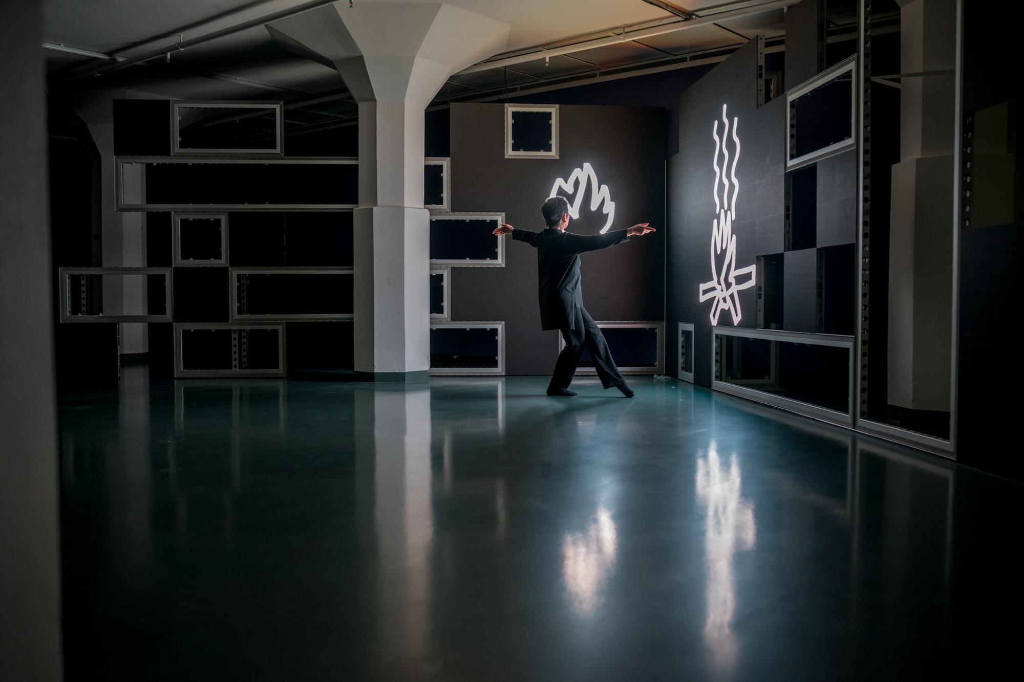 Danseres Muna Tseng voert een moderne dans uit in een donkere tentoonstellingsruimte, verlicht door neonachtige vlamvormen aan de muur.