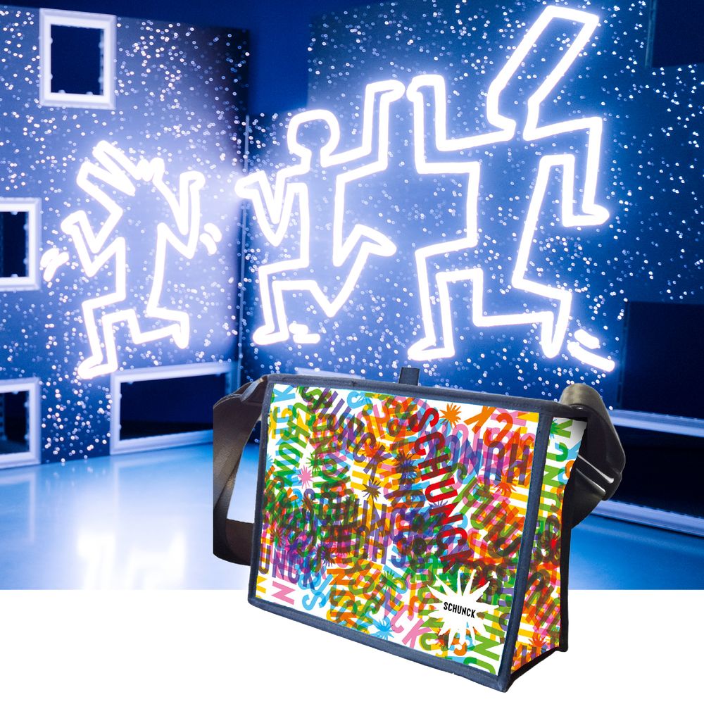 Kleurrijke museumtas met SCHUNCK-logo en speelse typografie staat voor een achtergrond met verlichte Keith Haring-figuren in een donkerblauwe ruimte vol lichtpuntjes.
