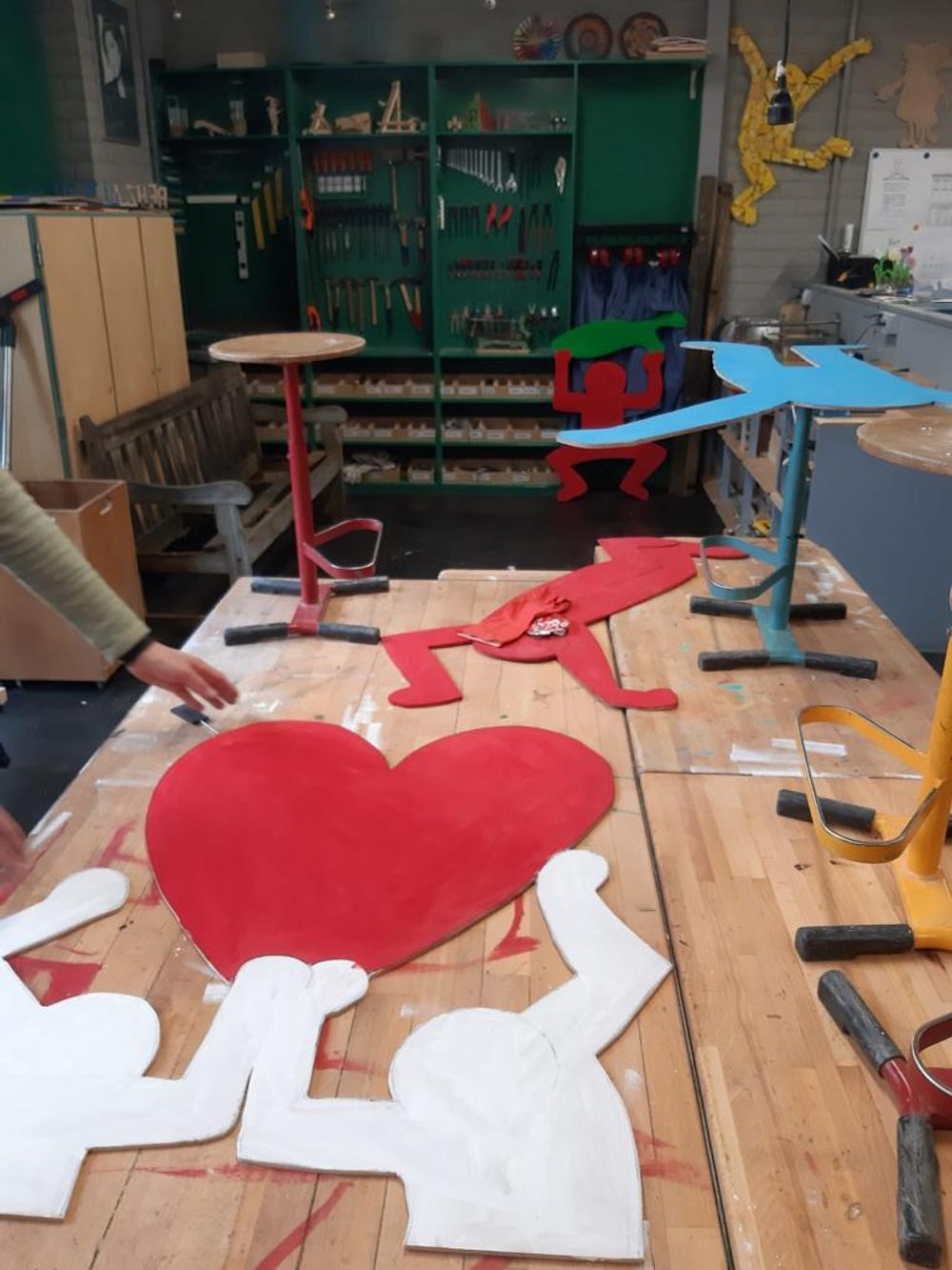 Werktafel in een creatieve werkplaats met gekleurde houten figuren in Keith Haring-stijl, waaronder een groot rood hart en dansende figuren. Op de achtergrond hangen gereedschappen aan een groene wand.