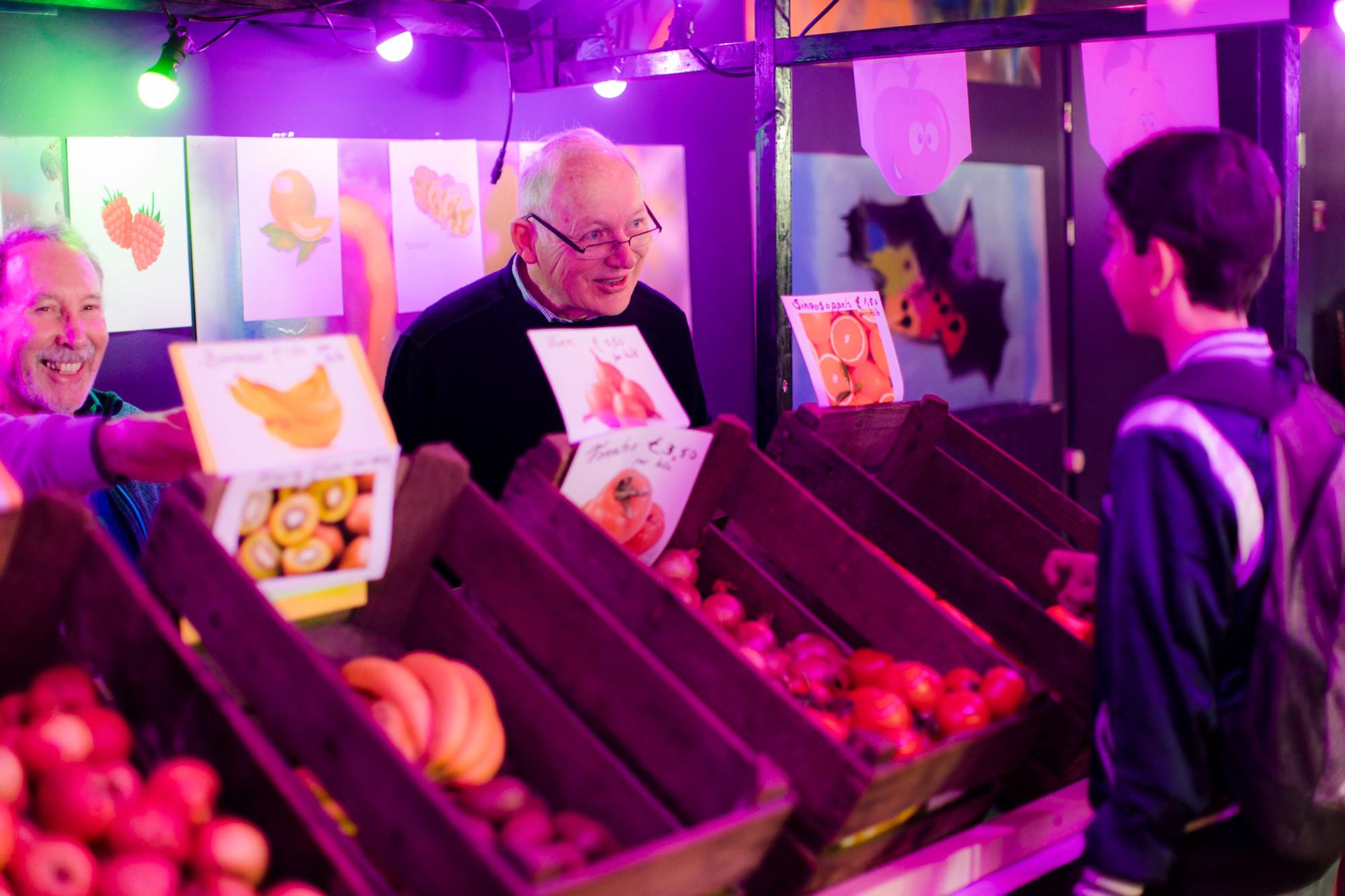 Twee mannen staan achter een marktkraam met fruit en praten met een jonge bezoeker tijdens het taaldorp.