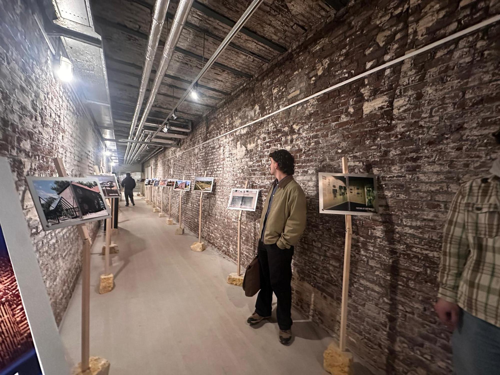 Lange, smalle tentoonstellingsruimte met bakstenen muren, waar foto’s op houten standaards langs de wand zijn opgesteld; een bezoeker staat stil en kijkt naar een van de werken.