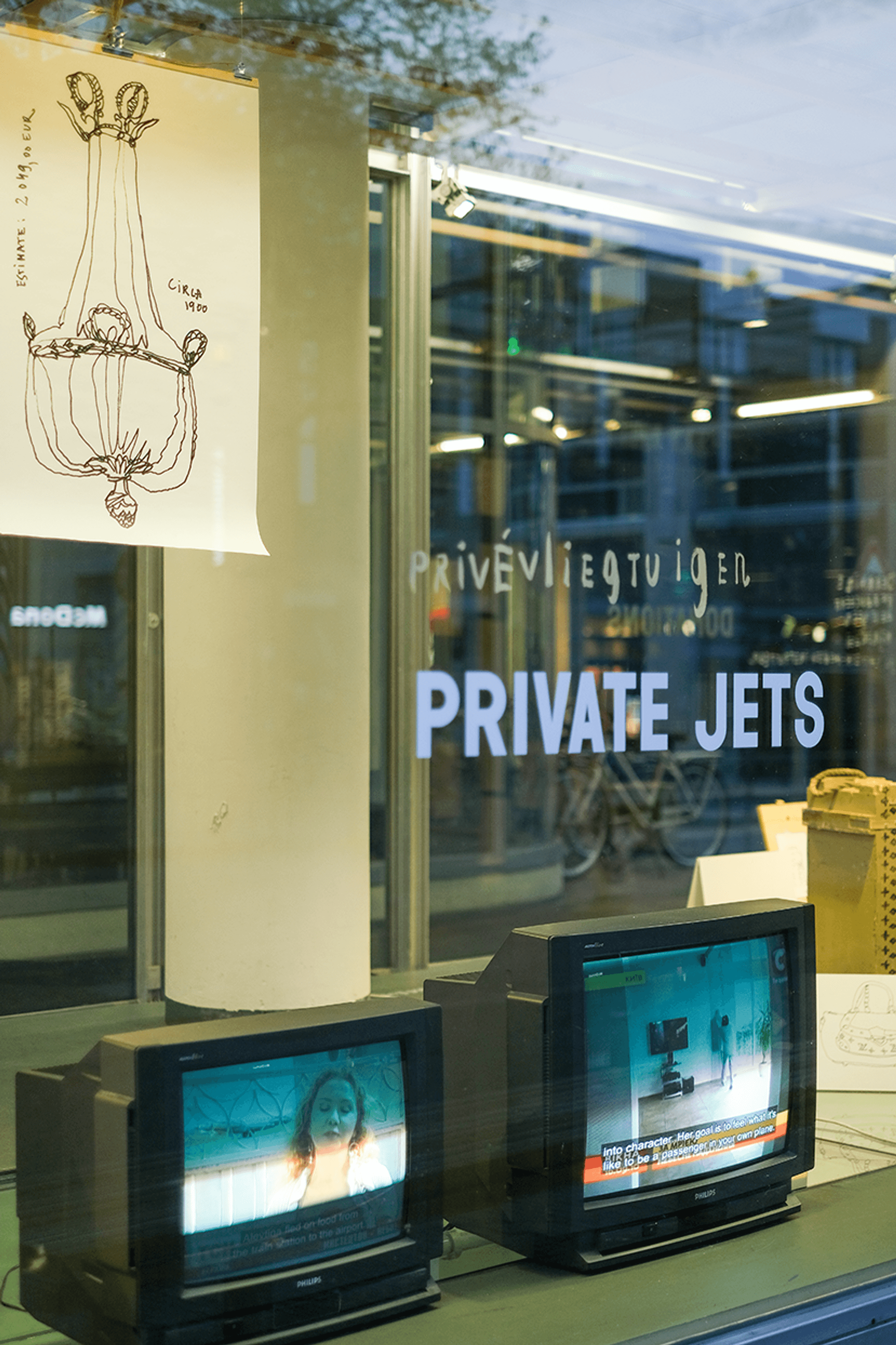 Twee ouderwetse televisies tonen videobeelden in een etalage; erboven hangt een tekening van een kroonluchter en op het raam staat de tekst 'Privévliegtuigen – Private Jets'.