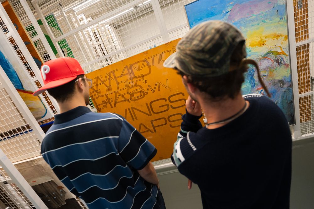 Twee mannen met een pet op, vanaf de rug gezien, kijkend naar verschillende kunstwerken.