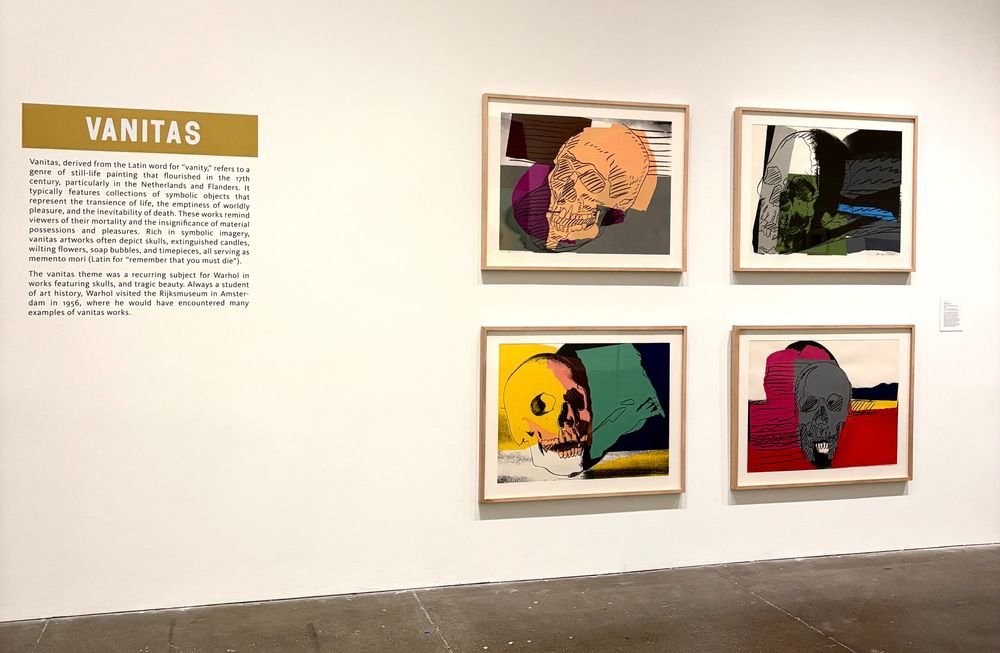 Een muur van de museumzaal van the Andy Warhol Museum in Pittsburgh met daarop een inleidende tekst over de kunststroming Vanitas en rechts daarvan 4 kunstwerken van Andy Warhol met daarop schedels.