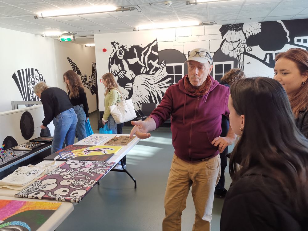 Man in paarse hoodie legt iets uit aan twee jonge vrouwen bij een tafel vol kleurrijke kunstwerken; op de achtergrond bekijken anderen kunst in een ruimte met zwart-witte muurschilderingen.