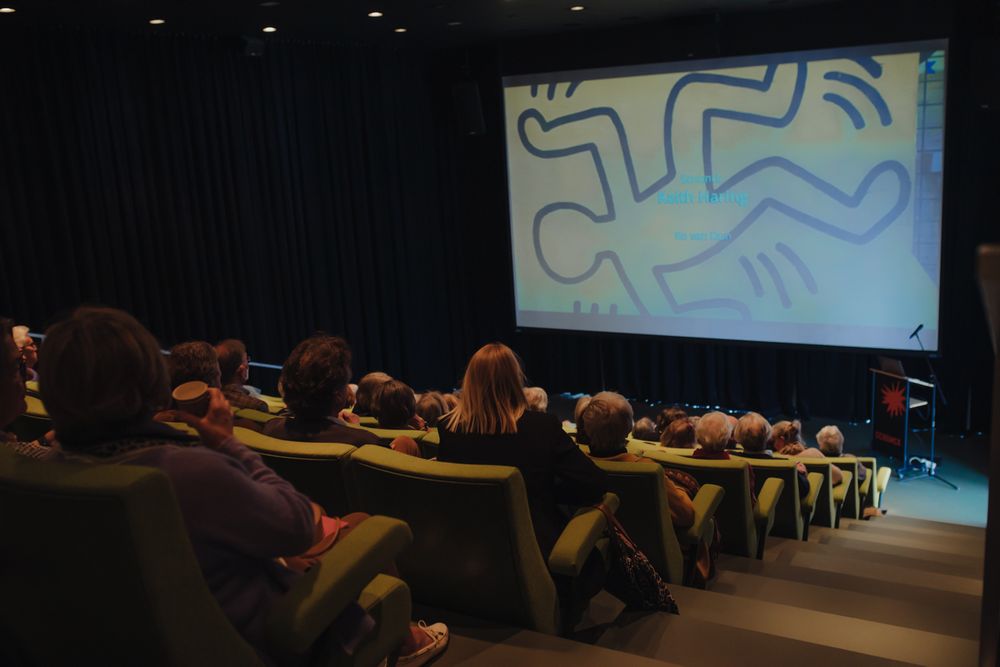 Bezoekers zitten in een theaterzaal met groene stoelen en kijken naar een presentatie over Keith Haring. Op het grote scherm is een illustratie in Haring-stijl te zien met de tekst “Keith Haring” en “Ko van Dun”. Rechts staat een katheder met het logo van SCHUNCK.
