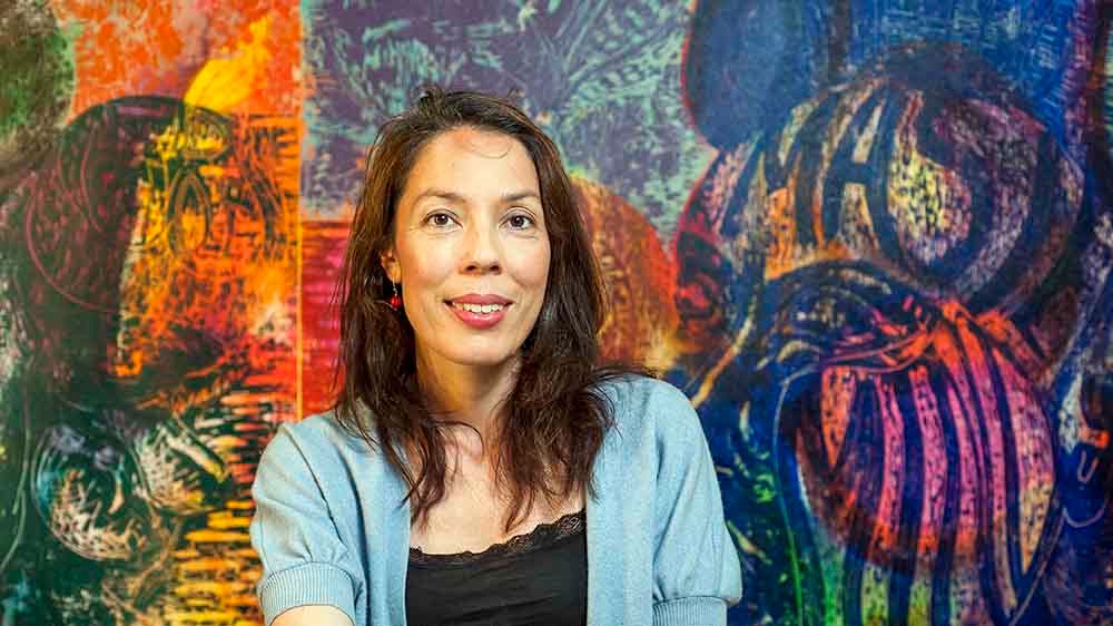 Een vrouw zit ontspannen voor een felgekleurde, expressieve achtergrond vol abstracte en figuratieve vormen. De combinatie van warme en koele kleuren geeft het beeld een levendige en energieke uitstraling.