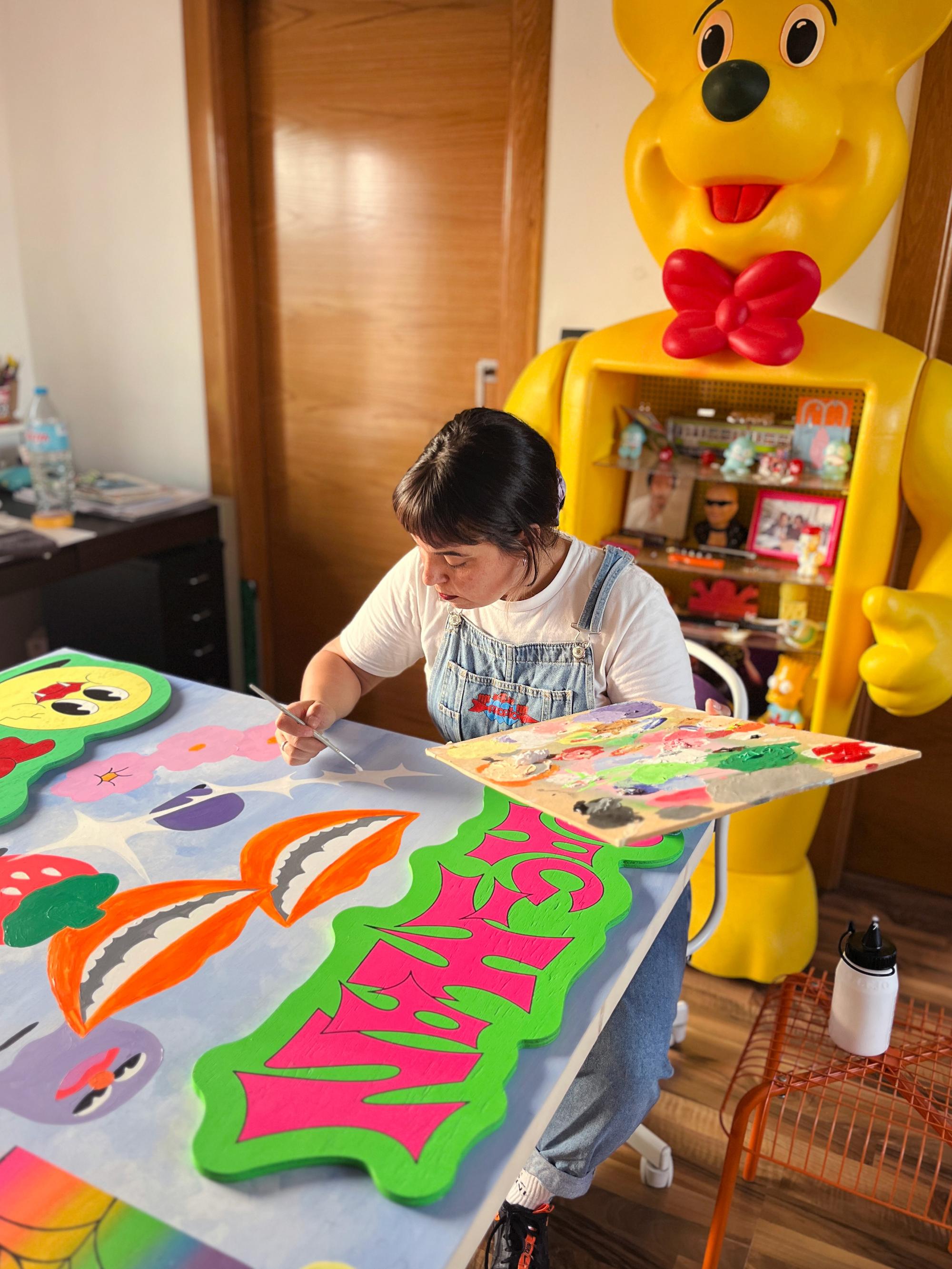 Kunstenaar Irene Fresh beschildert een groot kleurrijk werk in haar atelier, met op de achtergrond een grote gele berenkast vol verzamelobjecten.