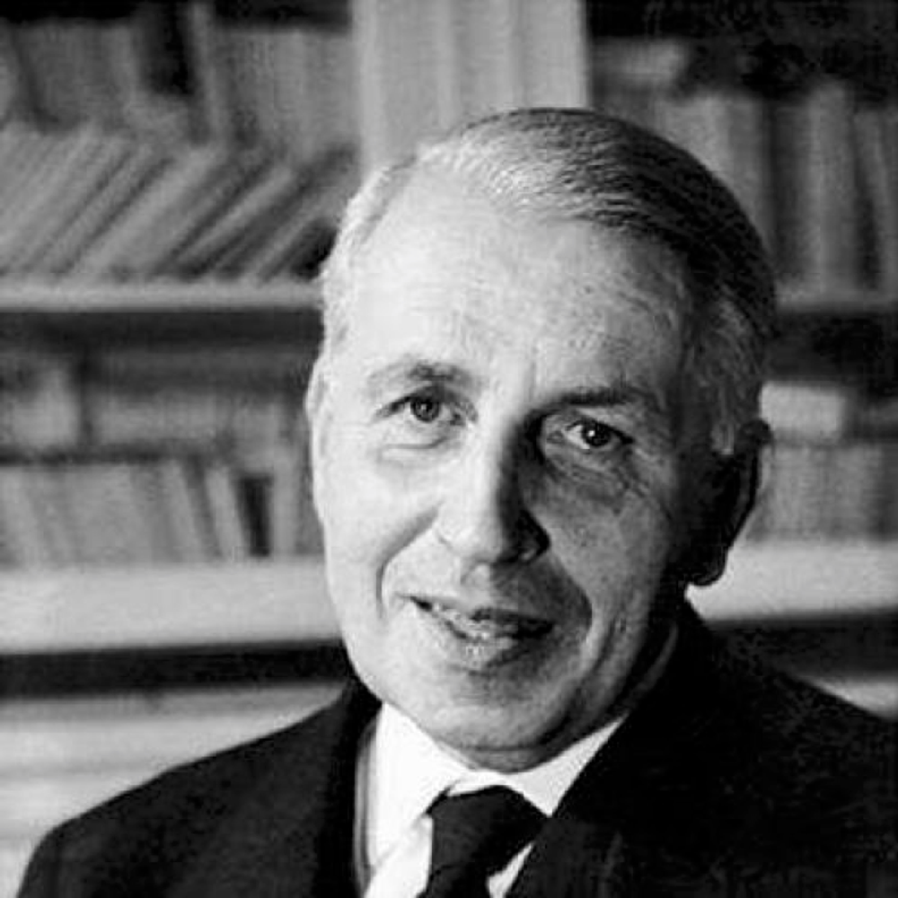 Zwart-witportret van Georges Battaile in formele kleding, gefotografeerd voor een boekenkast. De persoon kijkt recht in de camera, terwijl de achtergrond met boeken onscherp is weergegeven.