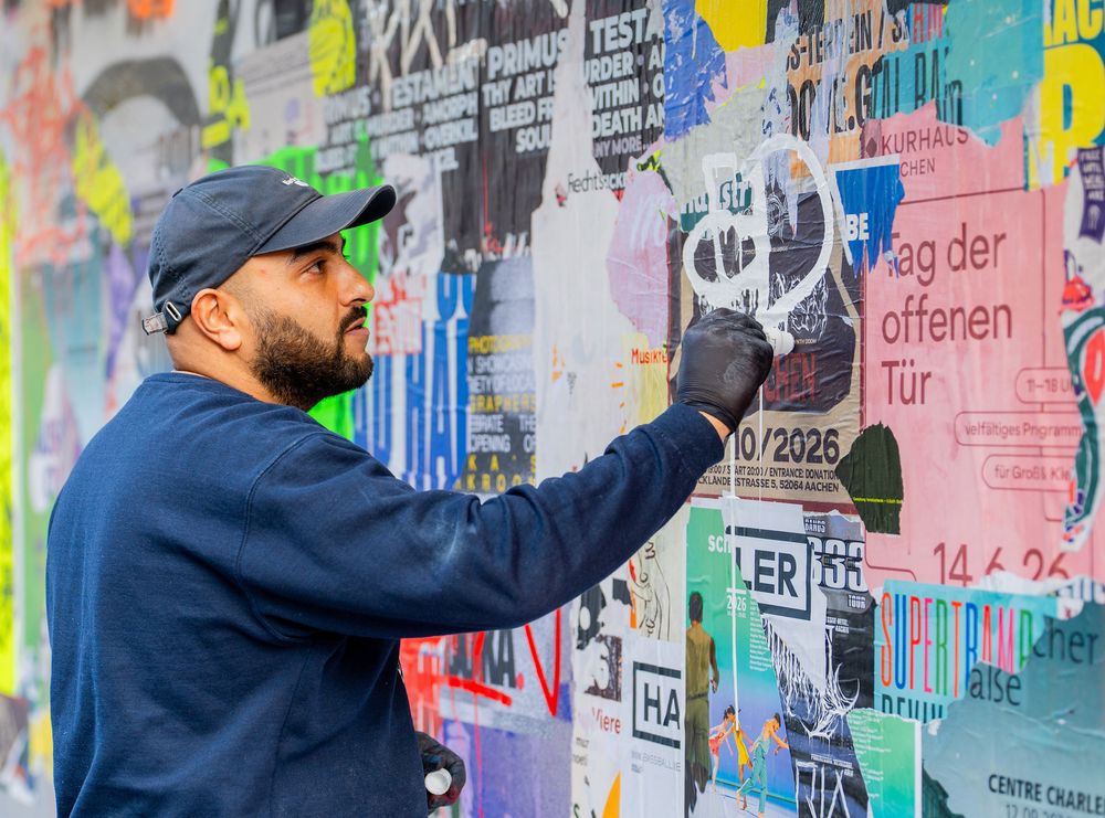 Close-up van kunstenaar Massoud Sakhi die met een zwarte handschoen witte graffiti aanbrengt op MODULE #6 bij het Glaspaleis in Heerlen. De muur is bedekt met kleurrijke posters, teksten en verflagen.