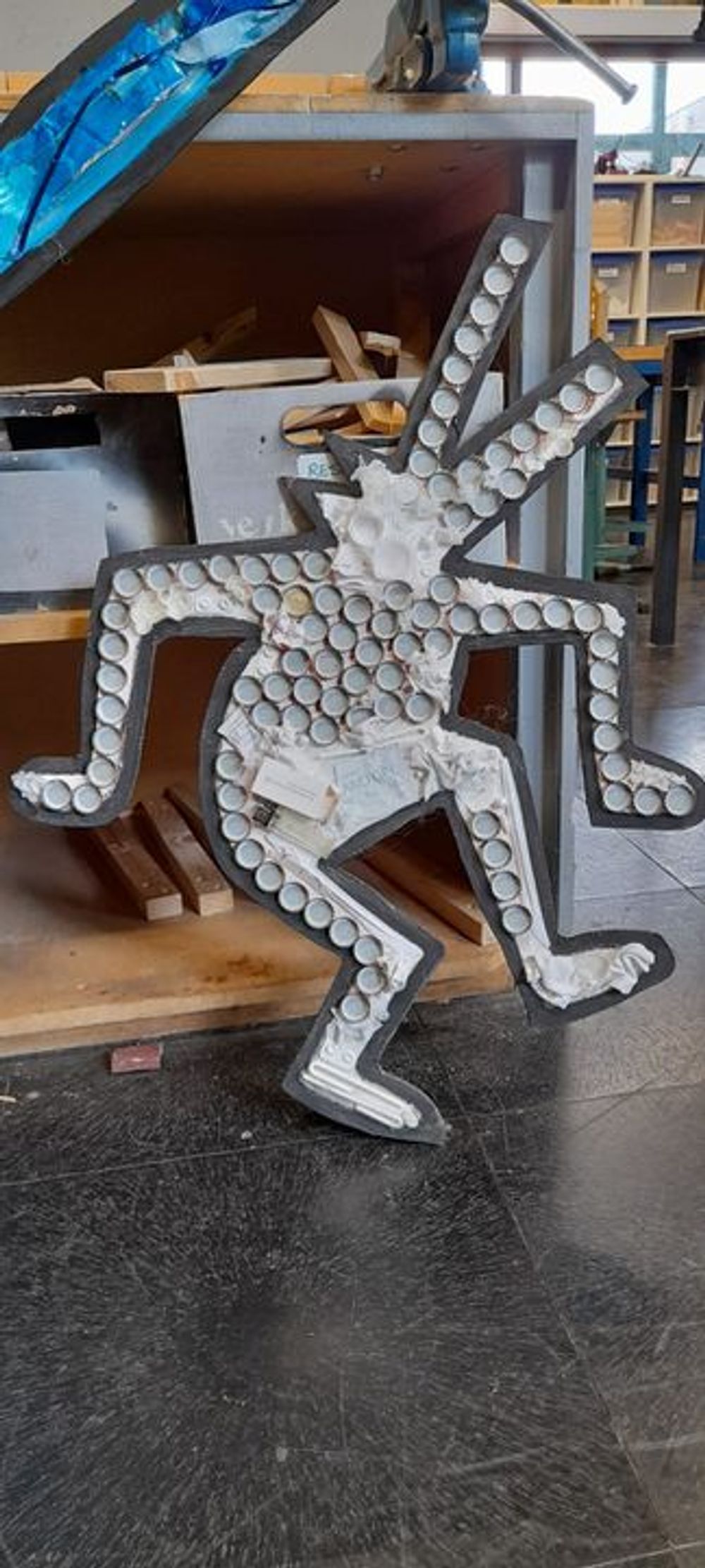 Mensfiguur gemaakt van cirkelvormige objecten: De figuur is samengesteld uit flessendoppen of vergelijkbare items, geplaatst op een zwart frame. De figuur staat op één been met het andere been omhoog en beide armen gebogen bij de ellebogen. Op de achtergrond zijn planken en verschillende items te zien in wat lijkt op een werkplaats of opslagruimte.


