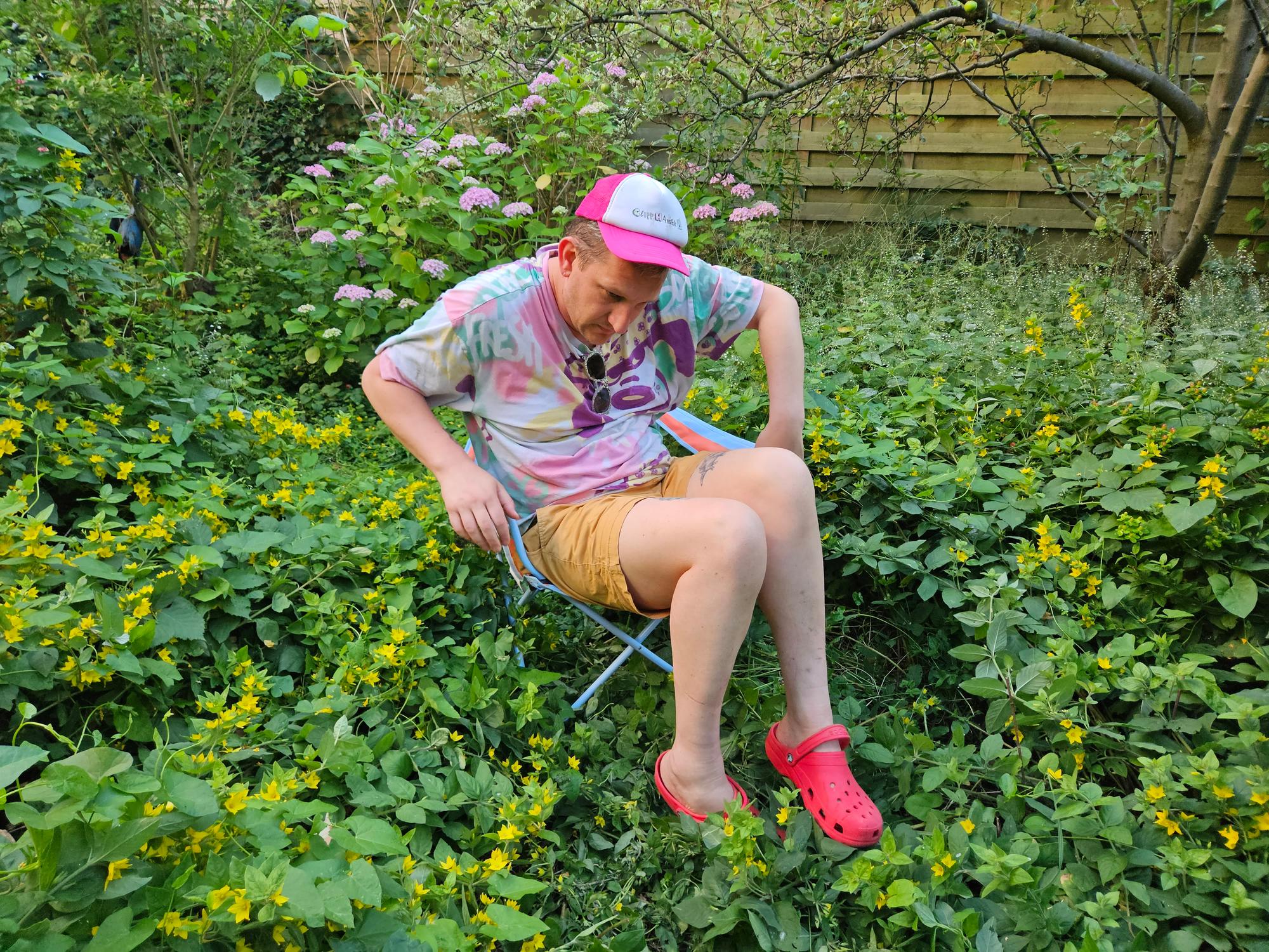 Maarten Bel, een man, zit ontspannen op een klapstoeltje in een weelderige tuin vol groene planten en gele bloemen. Hij draagt een kleurrijk T-shirt, beige korte broek, roze Crocs en een roze pet. Op de achtergrond zijn planten en een houten schutting te zien.