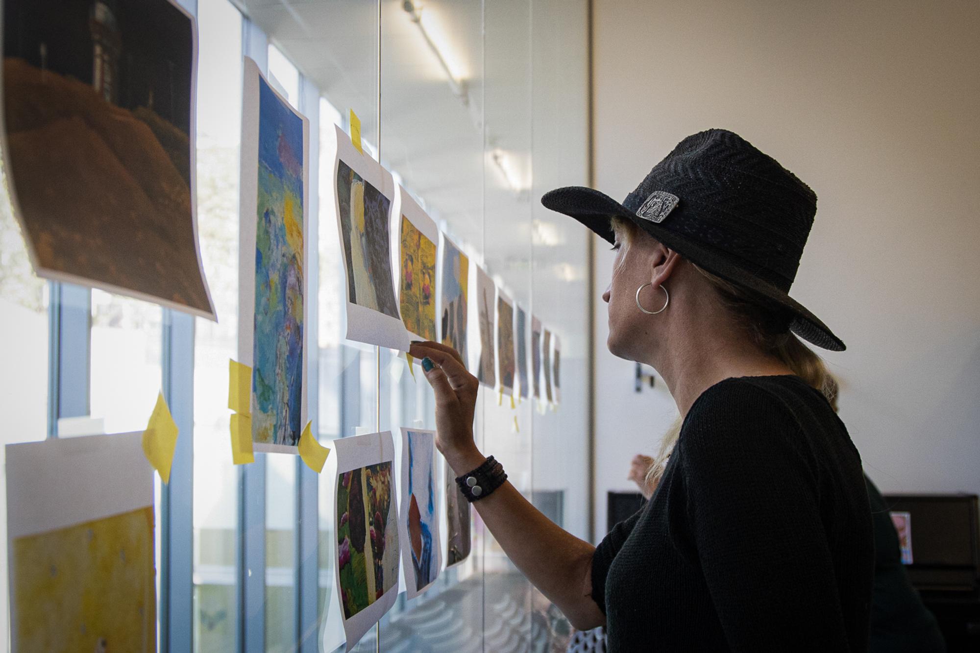 Een vrouw met een hoed bekijkt een selectie geprinte kunstwerken die op een glazen wand zijn geplakt.