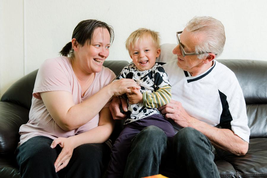 Vrouw en oudere man zitten op een zwarte bank met een vrolijk kind op schoot. Vrouw kietelt het kind. 