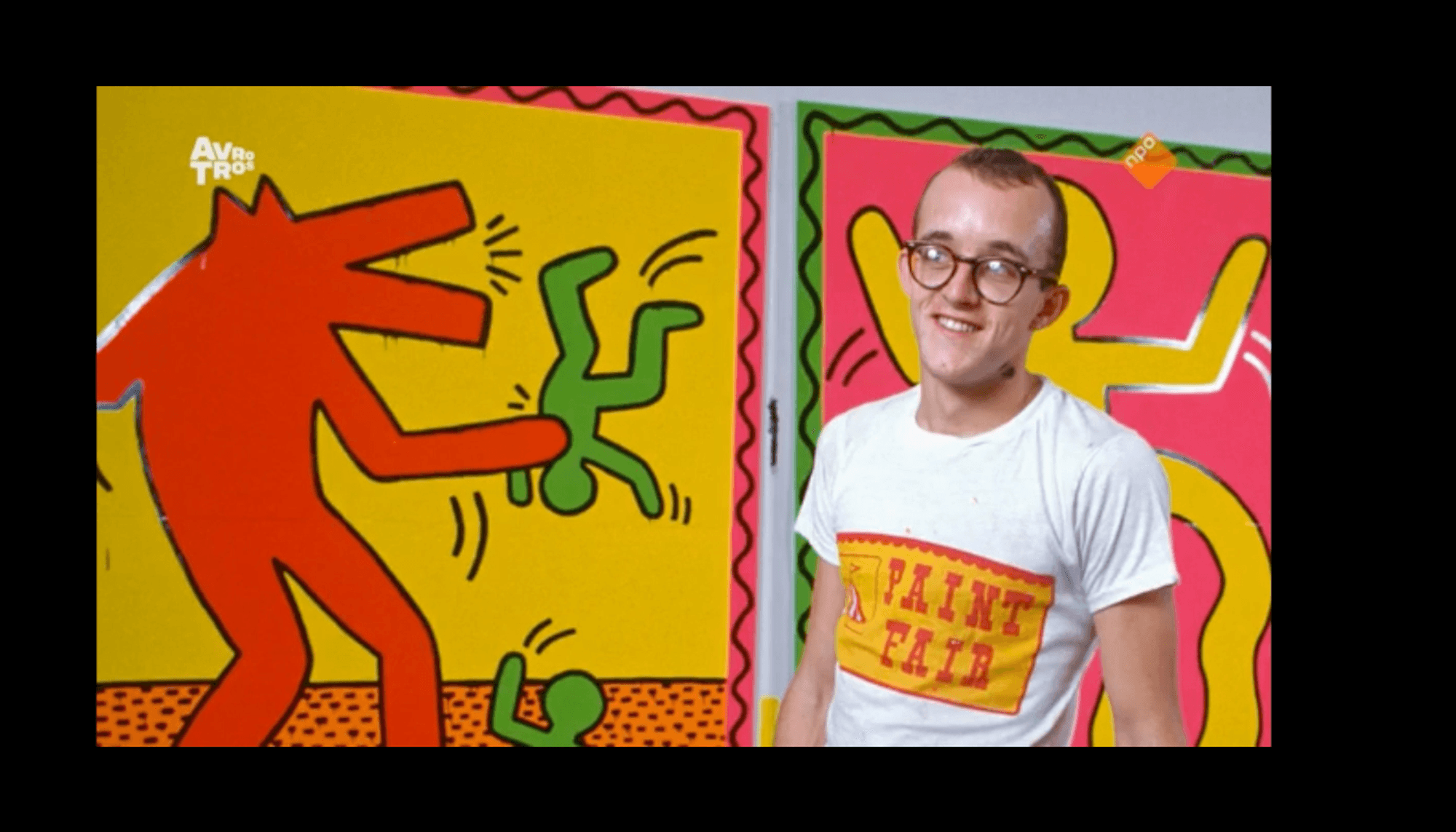 Een persoon staat voor twee kleurrijke kunstwerken. Het kunstwerk links toont een grote rode figuur die een kleinere groene figuur ondersteboven houdt, met een andere kleine groene figuur op de grond. Het kunstwerk rechts toont een grote gele figuur met opgeheven armen tegen een roze achtergrond. De persoon draagt een wit t-shirt met de tekst 'PAINT FAIR' in rood en geel.
