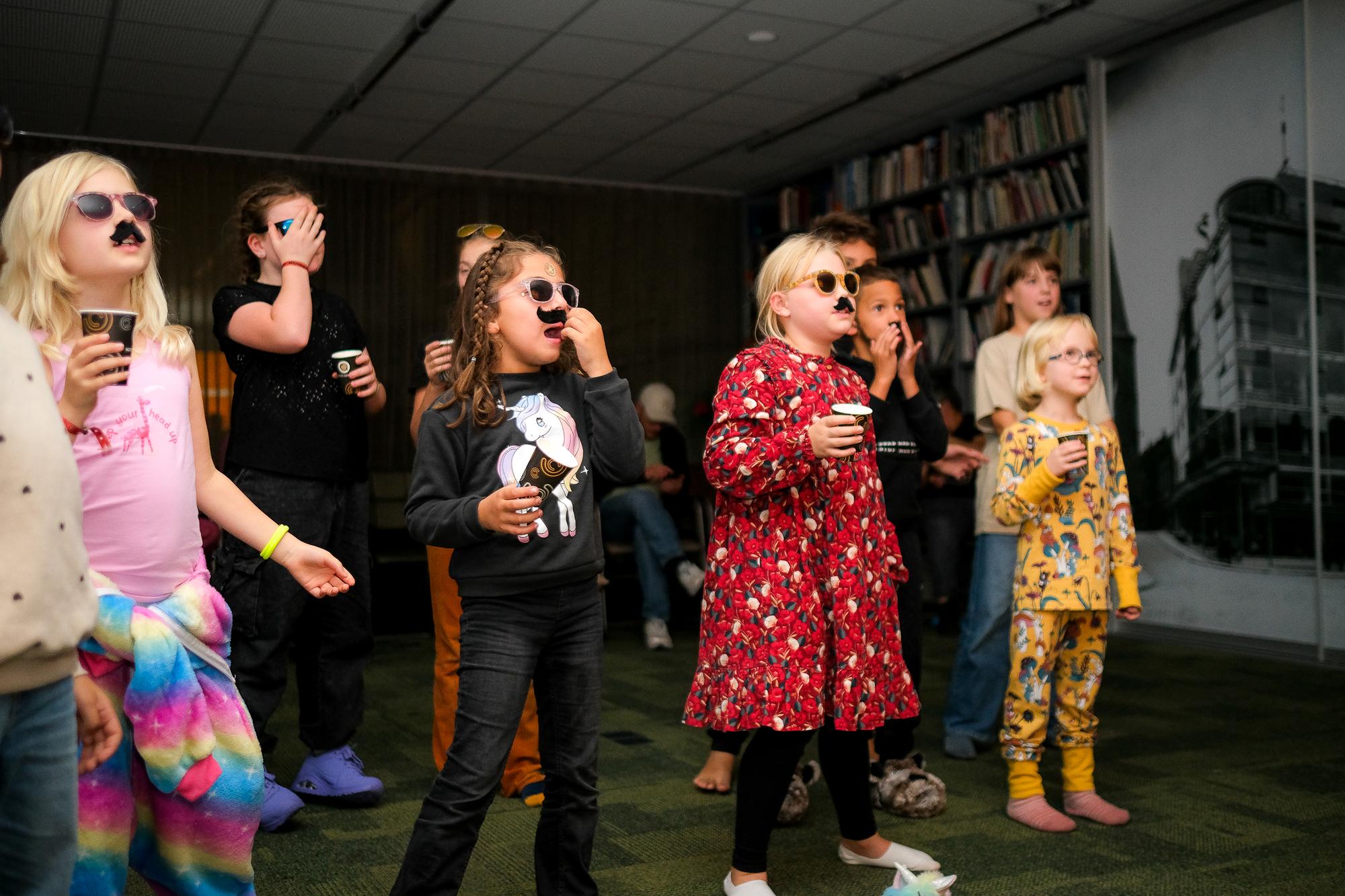 Groep kinderen staat in een ruimte met boekenkasten en draagt verkleedaccessoires zoals zonnebrillen en nep-snorr en terwijl ze drankjes vasthouden tijdens kamperen in de bieb, foto gemaakt door Eefje Pieters. 
