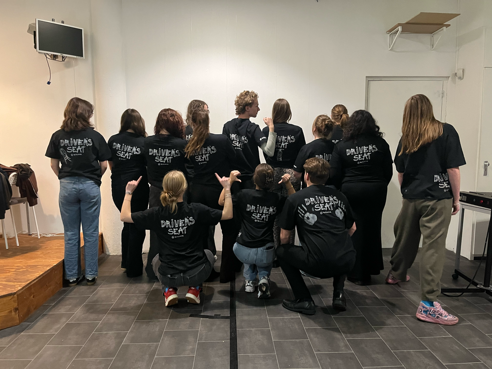 Groepsfoto van jongerengroep Driver's Seat met bedrukte t-shirts, waarvan alleen de rug van de jongeren zichtbaar is.