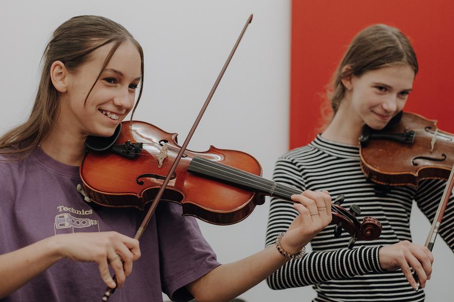 Twee personen bespelen violen in een binnenruimte. De persoon links draagt een paarse T-shirt met een grafische print en houdt de viool in speelpositie, terwijl de persoon rechts een gestreepte lange mouw draagt en eveneens een viool bespeelt. Op de achtergrond is een rode wand zichtbaar, wat contrasteert met de lichte muur.