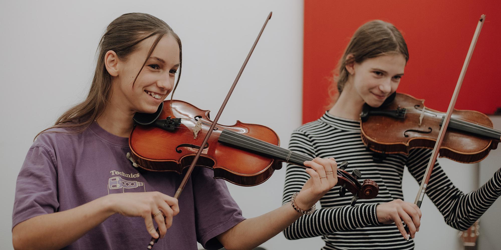 Twee personen bespelen violen in een binnenruimte. De persoon links draagt een paarse T-shirt met een grafische print en houdt de viool in speelpositie, terwijl de persoon rechts een gestreepte lange mouw draagt en eveneens een viool bespeelt. Op de achtergrond is een rode wand zichtbaar, wat contrasteert met de lichte muur.