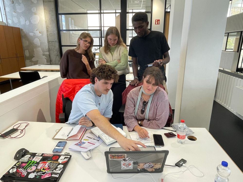 Vijf studenten bekijken samen ontwerpen op een laptop tijdens een werksessie. Twee studenten zitten aan tafel en drie anderen kijken over hun schouders mee.