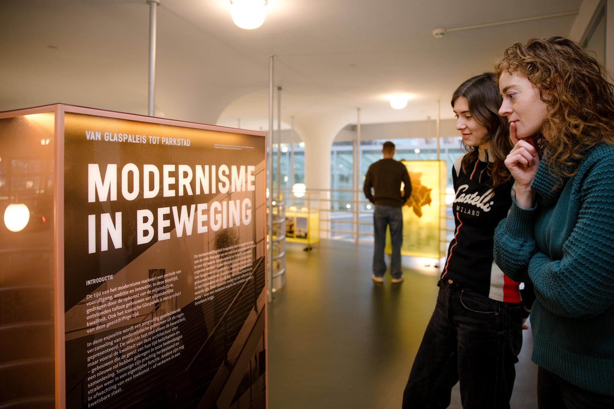 Twee bezoekers lezen een informatiepaneel met de titel 'Modernisme in Beweging' in een lichte tentoonstellingsruimte met witte zuilen.