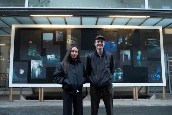 Twee jonge mensen staan buiten voor een groot kunstwerk met foto’s van nachtelijke stedelijke scènes in blauwe en groene tinten. Ze dragen donkere jassen en kijken in de camera, het kunstwerk hangt aan de gevel van een gebouw.