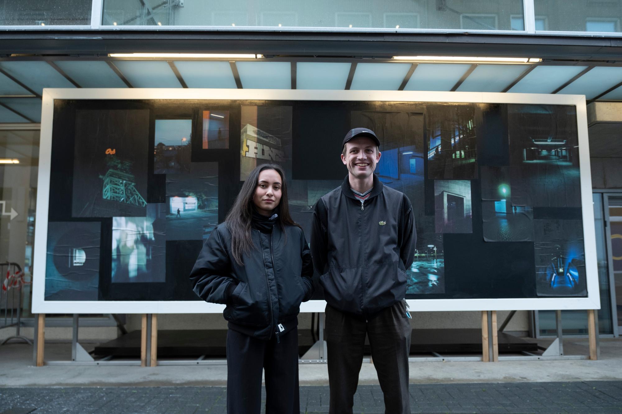 Twee jonge mensen staan buiten voor een groot kunstwerk met foto’s van nachtelijke stedelijke scènes in blauwe en groene tinten. Ze dragen donkere jassen en kijken in de camera, het kunstwerk hangt aan de gevel van een gebouw.